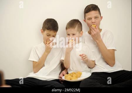 Glückliche, fröhliche Familie, die zusammen Muschis aß, öffneten ihren Mund und beißen köstliche Dessertkinder drei Söhne in weißen T-Shirts drei Kinder Stockfoto