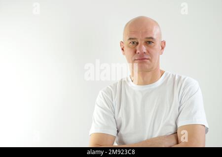 Ein Glatzkopf, der ein legeres weißes T-Shirt trägt, mit der Hand am Kinn und über Fragen nachdenklich ist. Lächelndes und rücksichtsvolles Gesicht. Zweifelhaftes Konzept. Ein erwachsener Glatzkopf legte seine Hände auf die Brust Stockfoto