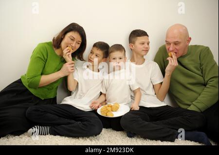 Fröhliche, fröhliche Familie, die zusammen Muschis isst, sie öffneten den Mund und beißen köstliche Desserts, Eltern in grünen Pullis, Kinder, drei Söhne in weißen T-Shirts, mutter, Vater und drei Kinder Stockfoto