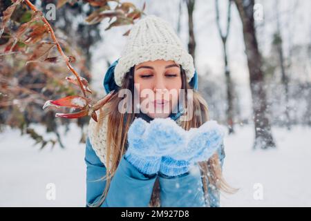 Glückliche junge Frau bläst Schnee aus den Händen, um Spaß im Winterwald zu haben. Mädchen mit blauem Mantel und Fäustlingen. Spielen im Freien bei kaltem Wetter Stockfoto