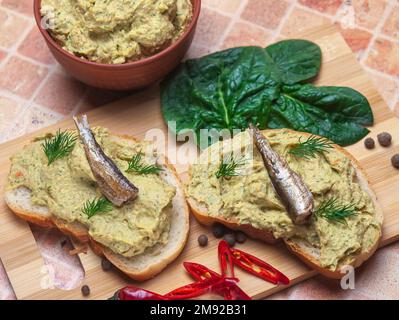 Sandwich mit Pastete und Sprotten. Sprottenpastete mit Spinat und Pfeffer. Sprotte in Öl, dänische Küche. Hintergrund zu Lebensmittelrezepten. Sandwich mit geräucherter Sprotte. Stockfoto