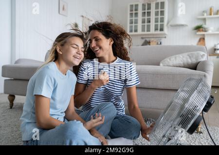Überglücklich Mutter und Teenager Tochter, die frische kalte Luft vom elektrischen Ventilator genossen, auf dem Boden sitzend Stockfoto