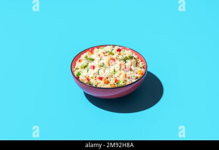 Schüssel mit gebratenem Reis und Gemüse minimalistisch auf einem bunten Tisch. Weißer Reis mit Karotten, rotem Pfeffer und grünen Bohnen in einer roten Schüssel. Stockfoto