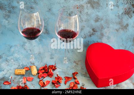 Zutaten zum Valentinstag (14. Oder 14. Februar) auf blauem Hintergrund: Flasche Rotwein, zwei Weingläser, herzförmige Geschenkbox, trockene Rosenblätter. Stockfoto