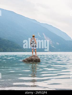 Junger Mann, der auf einem Felsen in der Mitte eines Sees steht, mit Bergen im Hintergrund. Sich jung zu fühlen, die Welt erkunden und bereisen zu wollen. Vertica Stockfoto