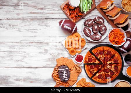 Superbowl- oder Fußballmotiv mit Food-Rand. Pizza, Hamburger, Flügel, Snacks und Beilagen. Blick von oben auf weißen Holzhintergrund. Stockfoto