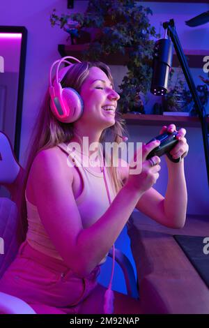 Seitenansicht einer positiven jungen Gamer-Spielerin in farbenfrohem Hemd und schnurlosem Headset, die neben dem Mikrofon sitzt, während sie streamt und Videospiele in Neon spielt Stockfoto