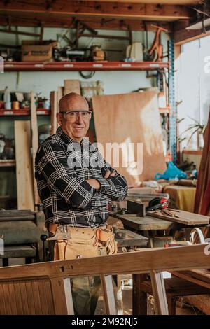 Fröhlicher alter Zimmermann im karierten Hemd und Schutzbrille, der lächelt und in die Kamera schaut, während er in Schreinern arbeitet Stockfoto