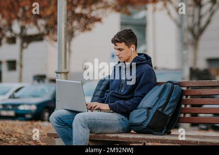 Fokussierter junger Mann in warmer Kleidung und Ohrhörern, der auf einer Holzbank mit Rucksack sitzt, während er im Herbstpark ein Notebook benutzt Stockfoto