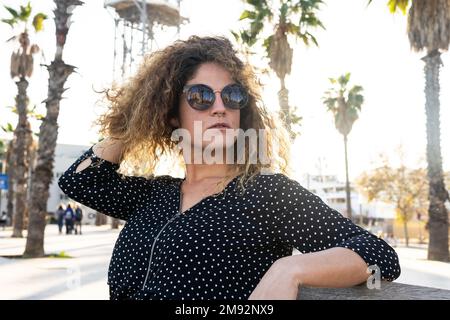 Frau mit Sonnenbrille und schwarzem gepunktetem Hemd, die an sonnigen Tagen am Wochenende auf einer Bank in der Nähe hoher Palmen ruht Stockfoto