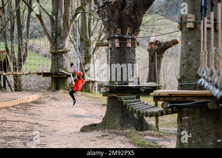 Seitenansicht eines nicht wiedererkennbaren Mädchens in warmen Kleidern, das auf einem Seil sitzt und Zeit im Seilpark im Herbstwald verbringt Stockfoto