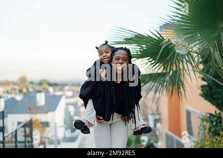 Eine afroamerikanische Frau mit Afro-Zöpfen, die in die Kamera schaut und einem lächelnden Mädchen draußen in der Nähe tropischer Pflanzen einen Huckepack gibt Stockfoto