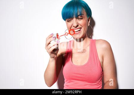 Positive junge Frau mit pinkfarbenem Oberteil und blauen Haaren, die Blasen bläst, während sie vor weißem Hintergrund steht und Spaß hat Stockfoto