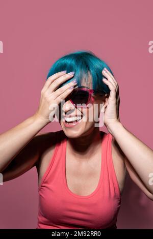 Glückliche junge Frau mit pinkfarbenem Oberteil und Sonnenbrille, die den Kopf berührt, während sie lächelt und im Studio steht Stockfoto