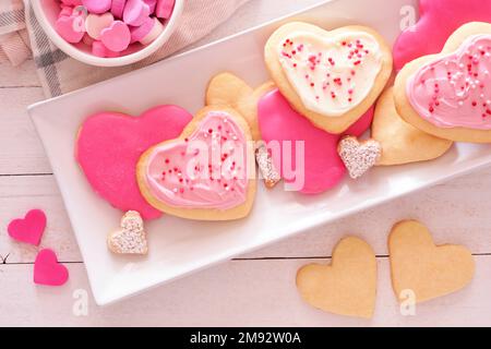 Herzförmige Valentinskekse mit pinkfarbenen und weißen Glasur und Streuseln. Blick von oben auf Servierteller mit weißem Holzhintergrund. Stockfoto