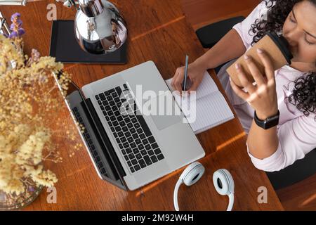 Draufsicht einer Freiberuflerin, die Kaffee aus einer Tasse zum Mitnehmen trinkt, während sie in der Nähe des Laptops auf einem Holztisch im Heimbüro in ihr Notizbuch schreibt Stockfoto