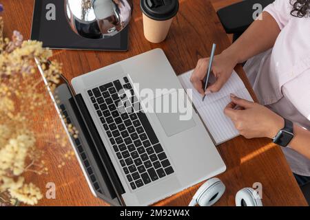Draufsicht einer freiberuflichen Freiberuflerin mit Tasse Kaffee zum Mitnehmen in Notizbuch neben Laptop auf Holztisch im Heimbüro Stockfoto