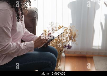Seitenansicht einer verkürzten, nicht erkennbaren Frau in Freizeitkleidung, die zu Hause mit einem Tablet auf dem Sofa sitzt Stockfoto