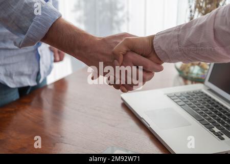 Ernten Sie anonyme männliche und weibliche Freiberufler, die sich nach einem erfolgreichen Geschäftsabschluss in der Nähe von Laptops und Tablets am Arbeitsplatz die Hände schütteln Stockfoto