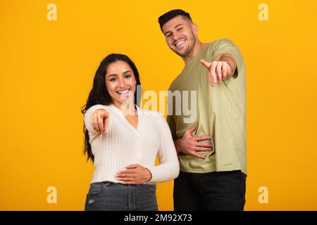 Lachender Millennials-araber und -Frau, ganz zwanglos, mit Fingern vor der Kamera, isoliert auf orangefarbenem Hintergrund Stockfoto