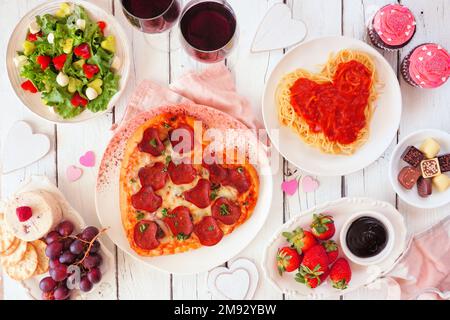 Hausgemachtes Abendessen am Valentinstag. Tischszene mit Blick von oben auf weißen Holzhintergrund. Herzförmige Pizza, Pasta, Wein, Käseplatte und Desserts. Stockfoto