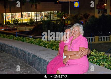 Zwei Frauen in leuchtend roten Kleidern stellen sich in der Nähe des Nachtladens auf der 5. Centennial Avenue, Puerto de Santiago, Teneriffa. Stockfoto