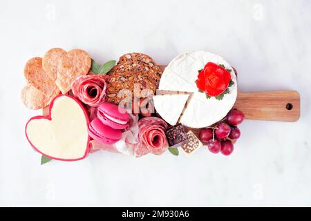 Wurstplatte am Valentinstag mit einer Auswahl an Käse, Vorspeisen und Obst. Blick von oben auf dunklem Holzhintergrund. Stockfoto