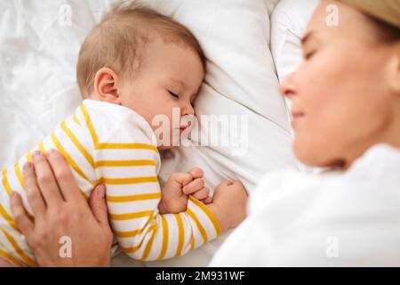 Liebende weiße Mutter umarmt schlafendes Baby, liegt im Bett, High Angle Shot, Fokus auf Kind Stockfoto