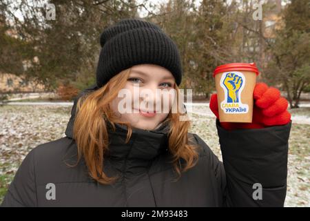 Das Mädchen hält in ihrer Hand eine Tasse Kaffee mit einem patriotischen Sticker. Übersetzung der Inschrift auf dem Aufkleber - "Ruhm der Ukraine" Stockfoto