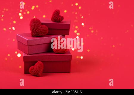 Drei rote Geschenkboxen mit gestrickten roten Herzen auf rotem Hintergrund mit goldenen festlichen Lichtern. Alles gute zum Valentinstag, Muttertag und Geburtstagsgrüße Stockfoto