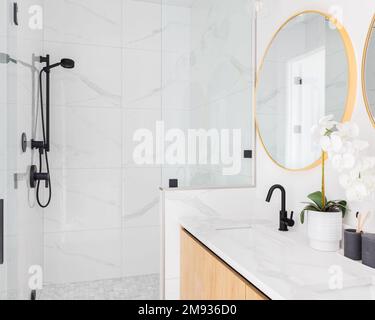 Luxuriöses Badezimmer mit schwimmendem Holzschrank, Marmorablagefläche, goldenen Spiegeln und marmorgefliester Duschkabine. Stockfoto