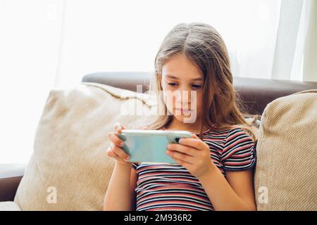 Süßes kleines Mädchen, das Technologie-Gerät benutzt, sitzt auf dem Sofa. Ein Mädchen, das am Telefon spielt. Stockfoto