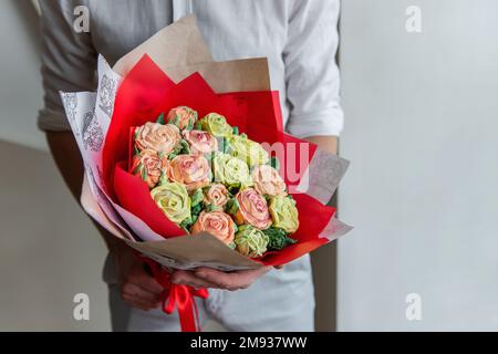 Ein gesichtsloser Mann hält einen Strauß süßer Marshmallow-Blumen in den Händen. Ein Geschenk in rotem Umschlag mit zarten Zuckerrosen auf hellem Hintergrund. Klein Stockfoto