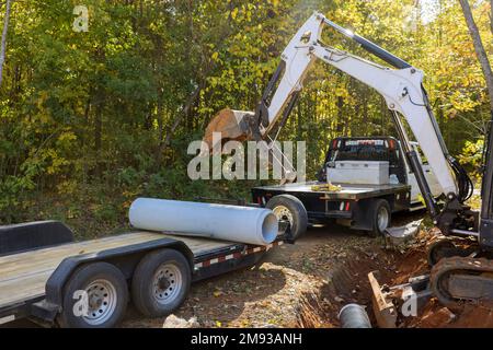 Arbeit eines Arbeiters, der den Traktor zum Anheben von Betonabwasserrohren bei der Bodenvorbereitung für unterirdische Installationsrohre verwendet Stockfoto