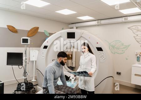 CT-Arzt konsultiert den Patienten und zeigt dem Patienten im Computertomographieraum den Röntgenkopf-Scan. Radiologe des CT-Scans zeigt dem Menschen eine Röntgenaufnahme des Gehirns Stockfoto