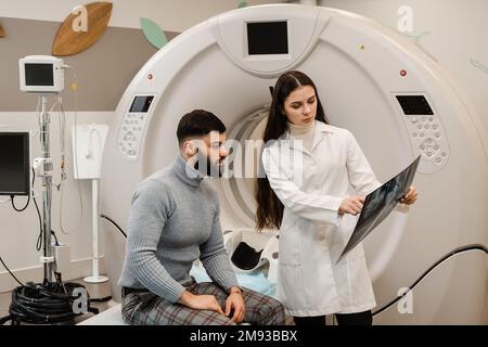 CT-Scan-Radiologe zeigt Röntgenaufnahmen des Gehirns einem Patienten im Computerscanraum. CT-Arzt konsultiert Patienten und zeigt Röntgenkopf-Scan zu Evidence Stockfoto