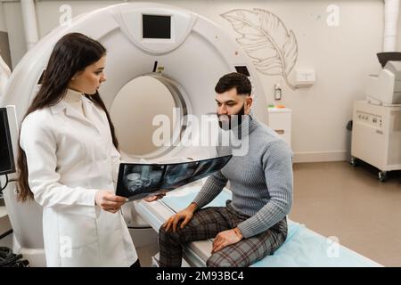 CT-Arzt, der den Patienten konsultiert und dem Patienten im Computertomographieraum das Thoraxröntgenbild zeigt. CT-Scan-Radiologe zeigt dem Patienten eine Röntgenaufnahme des Abdomens Stockfoto