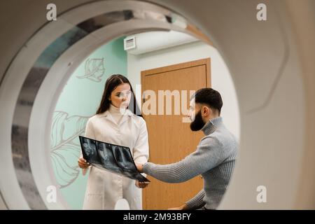 CT-Arzt, der den Patienten konsultiert und dem Patienten im Computertomographieraum das Thoraxröntgenbild zeigt. CT-Scan-Radiologe zeigt dem Patienten eine Röntgenaufnahme des Abdomens Stockfoto
