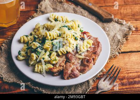Schweinehals-Steak und cremige Pasta mit Spinat und Thymian Garniert mit geriebenem Parmesankäse auf weißem Teller Stockfoto