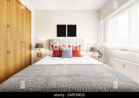 Doppelzimmer mit einem Bett mit grauem Kopfteil, zwei Lampen auf den Nachttischen, einem Fenster mit halbtransparentem Rollo und einem Kleiderschrank mit Eichentür Stockfoto