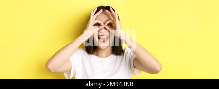 Nahaufnahme der lustigen asiatischen Mädchen durch Hand Fernglas und lächelnd, stehend in T-Shirt vor gelbem Hintergrund Stockfoto