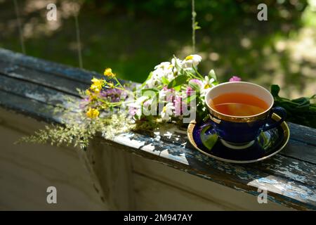 Tasse Tee und Blumenstrauß Kamillenblumen und Wildblumen auf Holztisch im Garten, verschwommener Hintergrund, weicher Fokus Stockfoto