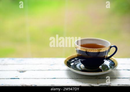 Eine Tasse Tee auf der Terrasse im Sommergarten. Schwarzer oder Kräutertee in einem wunderschönen blauen Becher auf einem grünen, verschwommenen Hintergrund Stockfoto