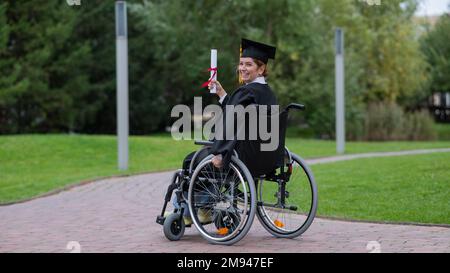 Glückliche weiße Frau im Rollstuhl dreht sich um und hält ihr Diplom draußen. Stockfoto