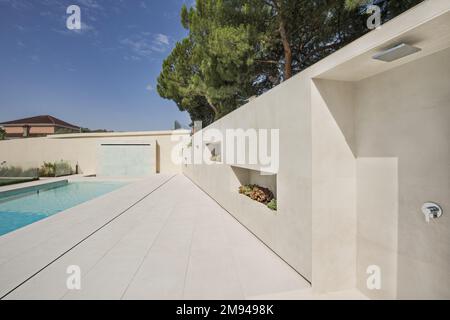 Duschbereich eines Swimmingpools mit weißen Marmorfliesen und Wand aus demselben Material mit Nischen für Pflanzen Stockfoto