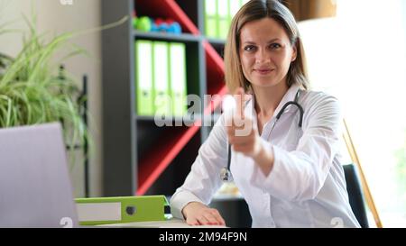 Eine Ärztin sitzt an ihrem Schreibtisch in der Klinik und zeigt Daumen nach oben. Stockfoto