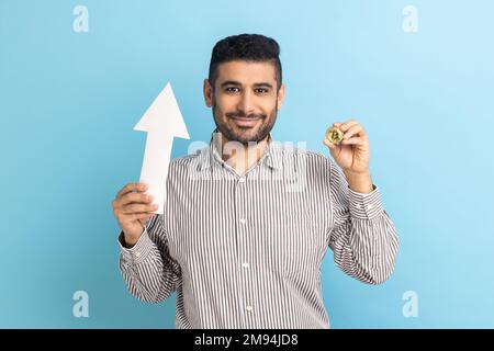 Porträt eines jungen erwachsenen attraktiven Geschäftsmannes, der eine Zunahme von Bitcoin zeigt, positive Emotionen ausdrückt und ein gestreiftes Hemd trägt. Innenstudio, isoliert auf blauem Hintergrund. Stockfoto