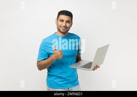 Porträt eines glücklichen, entzückten, attraktiven, unrasierten Mannes, der ein blaues T-Shirt trägt, den Laptop in den Händen hält, online arbeitet und Daumen nach oben zeigt. Studioaufnahmen im Innenbereich isoliert auf grauem Hintergrund. Stockfoto