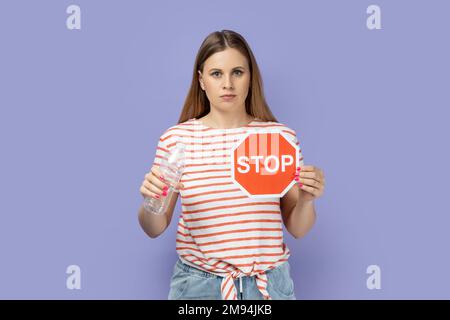 Porträt einer ernsthaften, streng herrisch jungen blonden Frau, die gestreiftes T-Shirt trägt, mit rotem Stoppschild und einer Plastikflasche in den Händen. Studio-Aufnahme im Innenbereich isoliert auf lila Hintergrund. Stockfoto