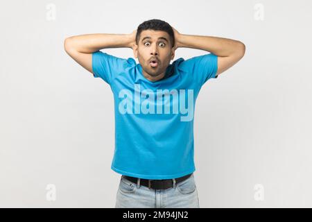 Portrait eines schockierten, überraschten Mannes, der ein blaues T-Shirt trägt und mit großen Augen in die Kamera schaut, die Hände hinter dem Kopf hält, etwas sieht, was erstaunt ist. Studioaufnahmen im Innenbereich isoliert auf grauem Hintergrund. Stockfoto